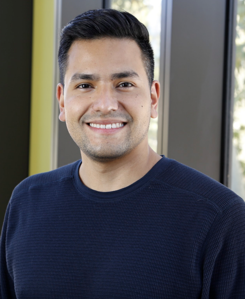 Oscar Leong smiling in a dark sweater in an indoor portrait with large windows behind him.